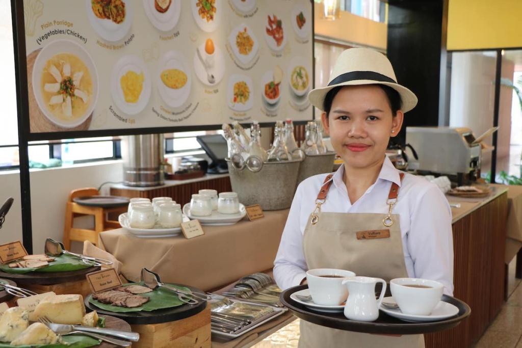 A female server in a hat and apron holding a tray with two cups of coffee at The Flock Cafe at The Aviary Hotel Siem Reap